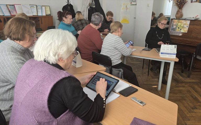 Mokymų dalyviai prie stalų mokosi naudotis planšetiniais kompiuteriais