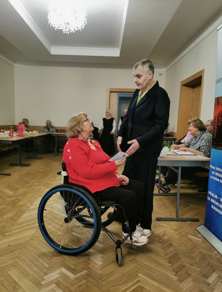Darbuotoja draugijos salėje kalbasi su moterimi neįgaliojo vežimėlyje